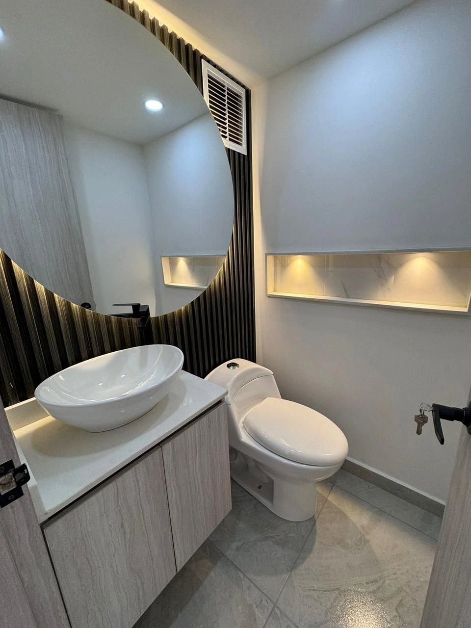 Baño con espejo circular, muro listonado y nicho iluminado